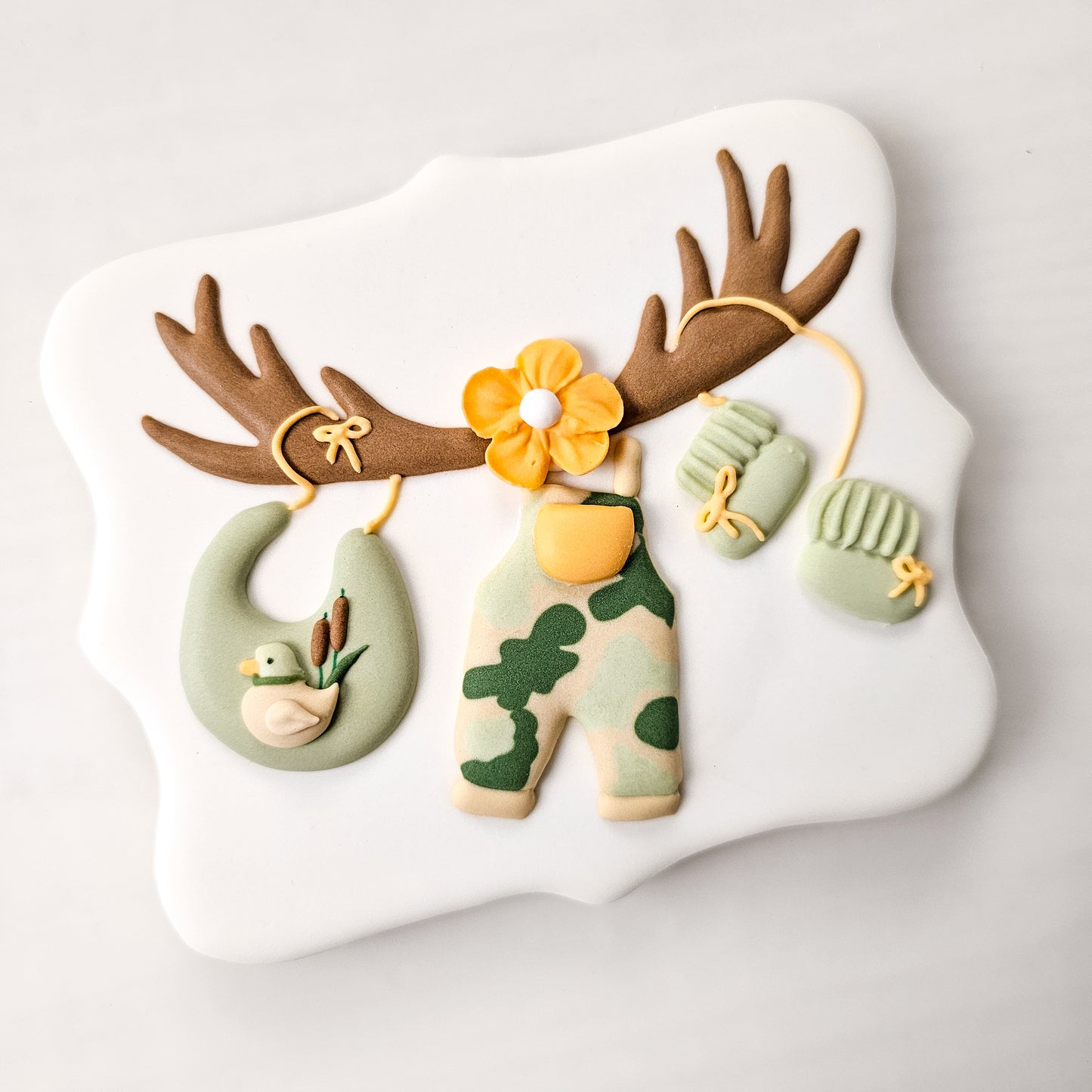 Decorative sugar cookie with a deer antler clothesline line with a bib with a duck on it, baby overalls, and baby booties royal icing transfers in a camouflage theme