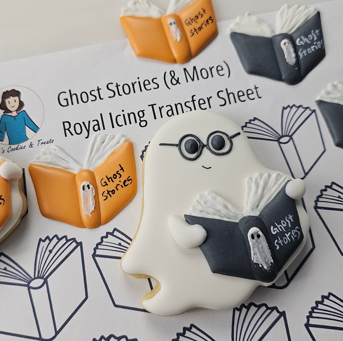 Decorated ghost-shaped iced cookie reading a royal icing transfer book, arranged on top of a printed transfer sheet titled 'Ghost Stories (& More) Royal Icing Transfer Sheet'.