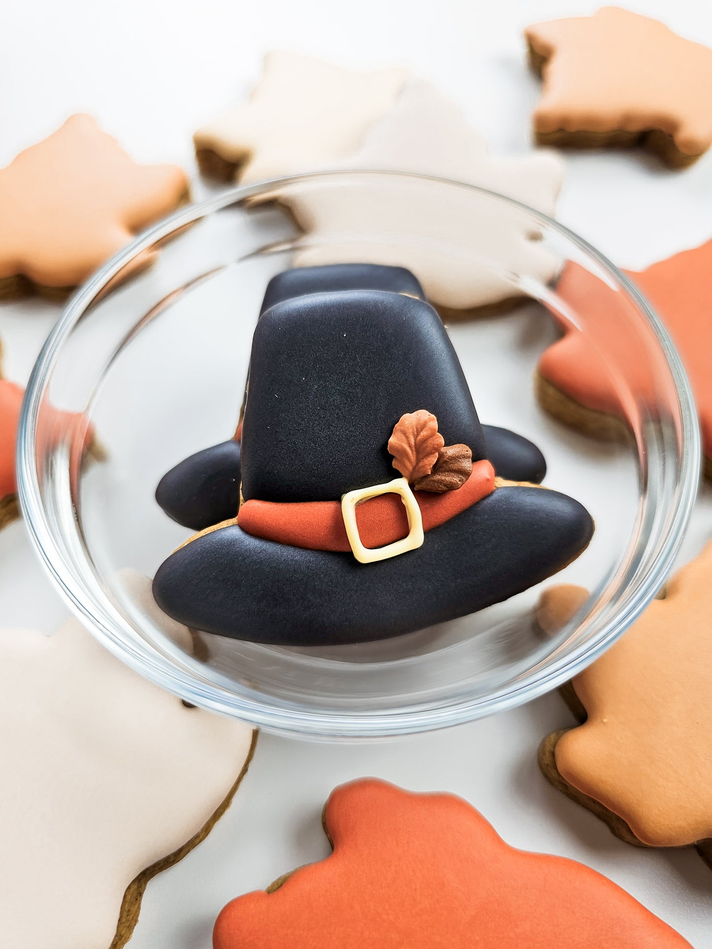 Decorative black pilgrim hat cookie on a clear plate with other decorated cookies in the background.