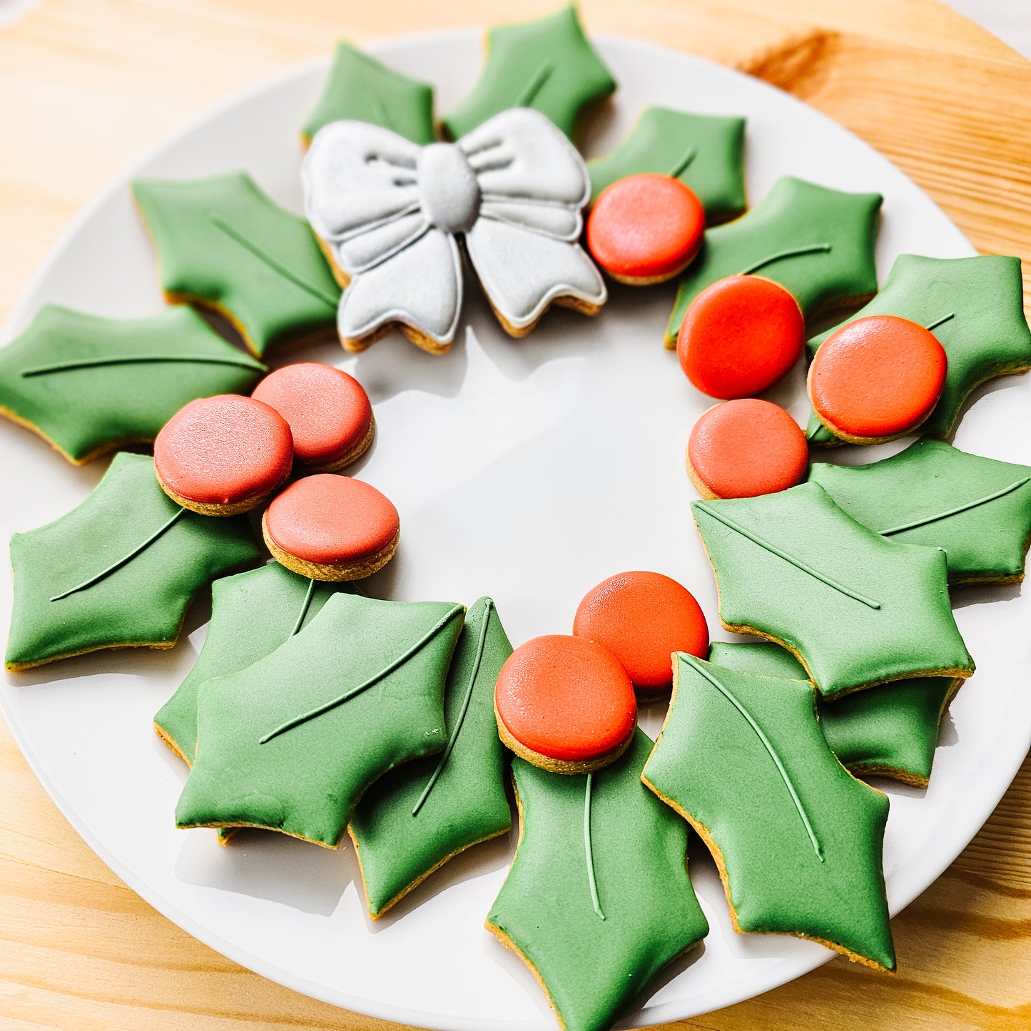 Decorative sugar cookies shaped like a Christmas wreath with green leaves, red berries, and a silver bow on a white plate.