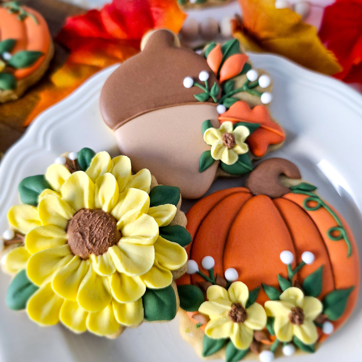 Three decorated sugar cookies shaped like an acorn, sunflower, and pumpkin, arranged on a white plate with autumn leaves in the background.