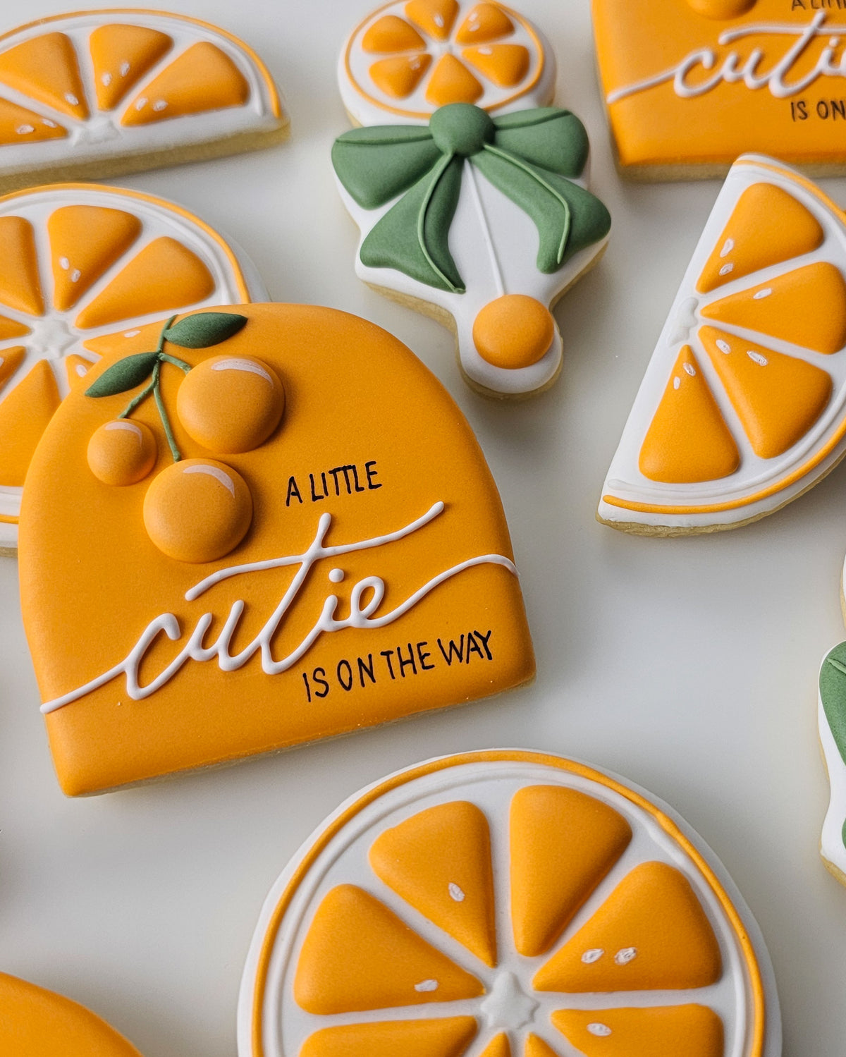 A close-up of decorated sugar cookies shaped like orange slices and a citrus slice with the phrase “A LITTLE CUTIE IS ON THE WAY” in white icing, arranged on a white surface.