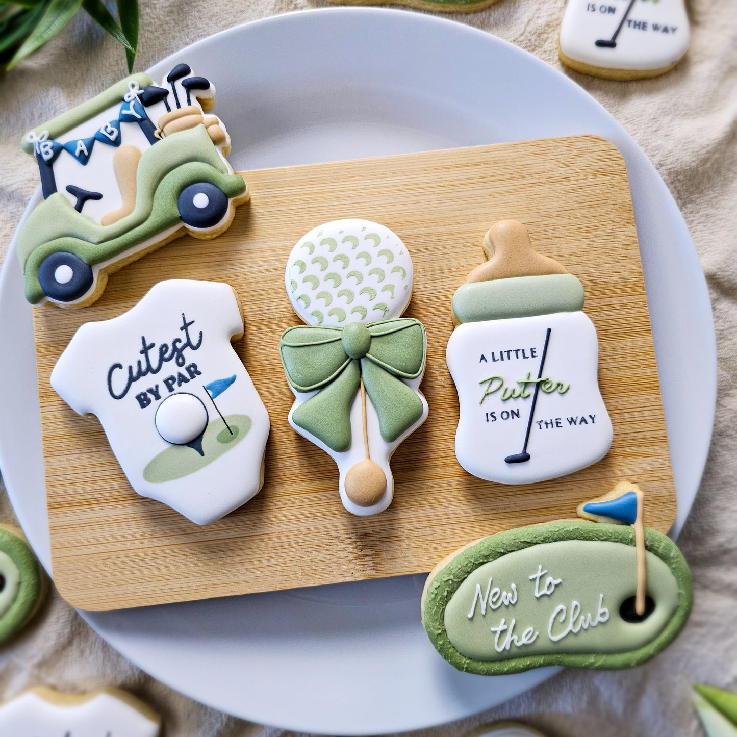 Decorative golf-themed sugar cookies shaped like a baby bottle, onesie, rattle, putting green, and golf cart on a wooden board.