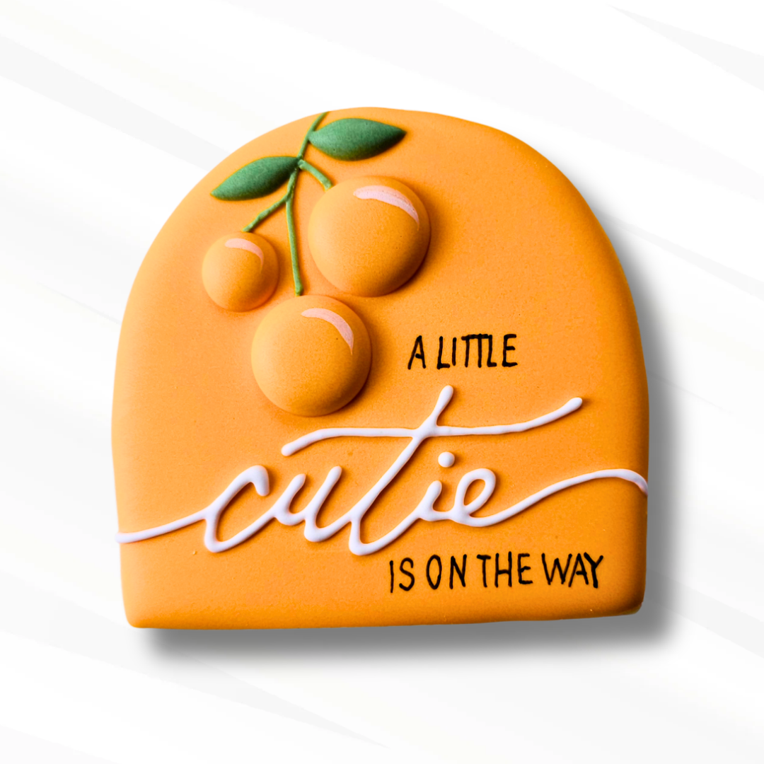 An orange-shaped decorative plaque cookie with a 3D fruit design and the text 'A LITTLE CUTIE IS ON THE WAY' on a white background.