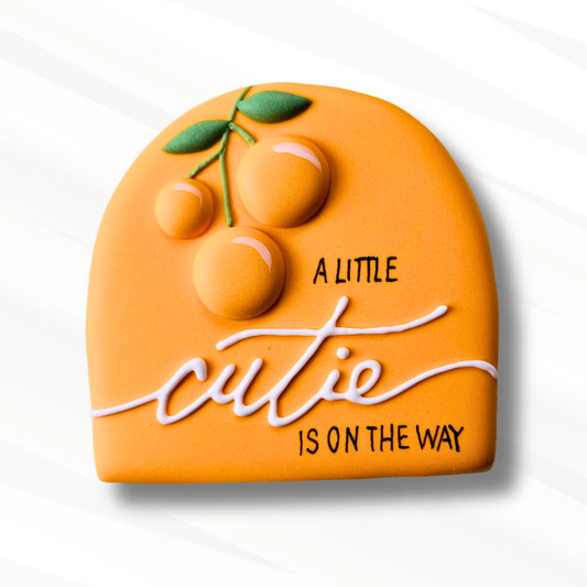 An orange-shaped decorative plaque cookie with a 3D fruit design and the text 'A LITTLE CUTIE IS ON THE WAY' on a white background.