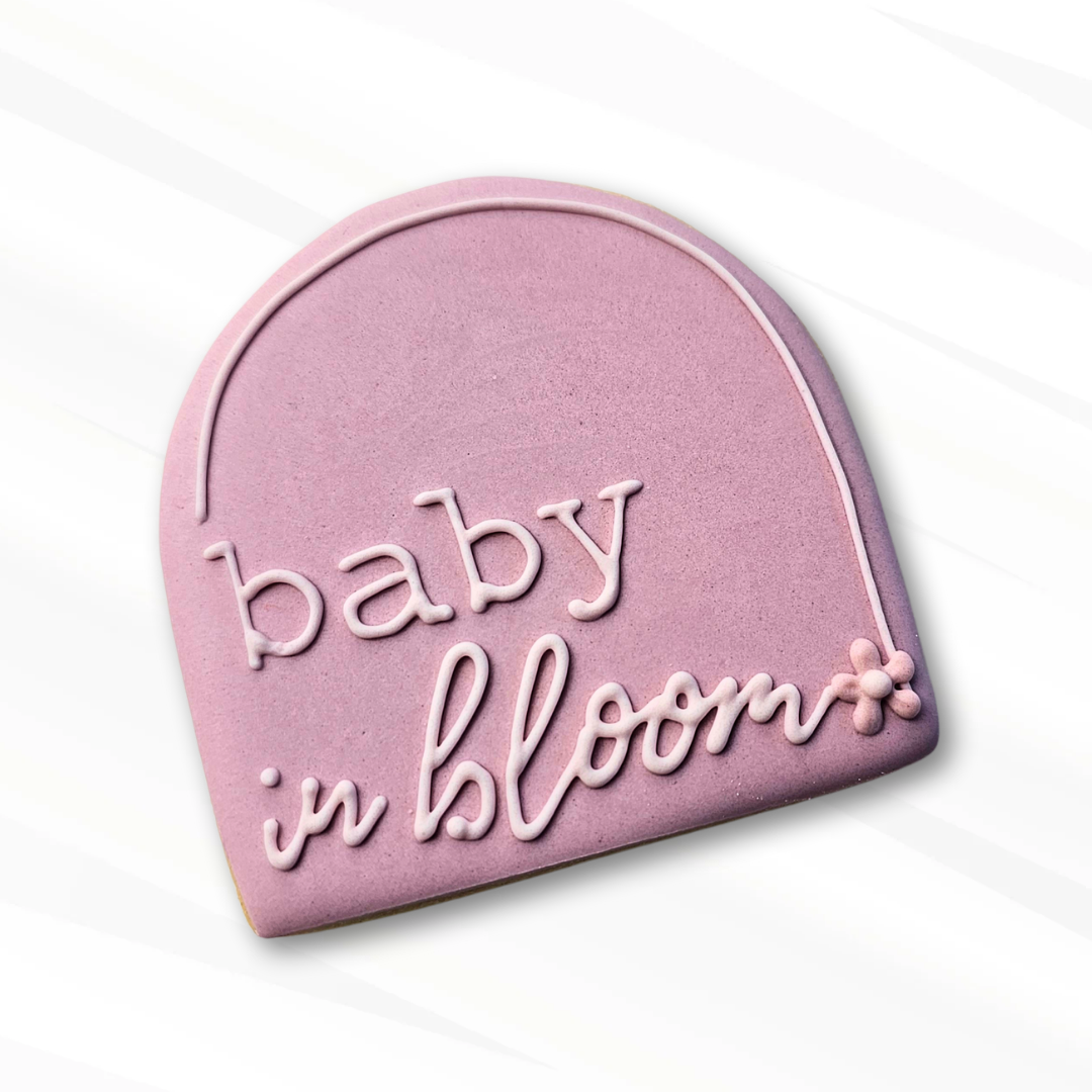 Pink baby shower arch shaped sugar cookie with 'baby in bloom' text on a white background