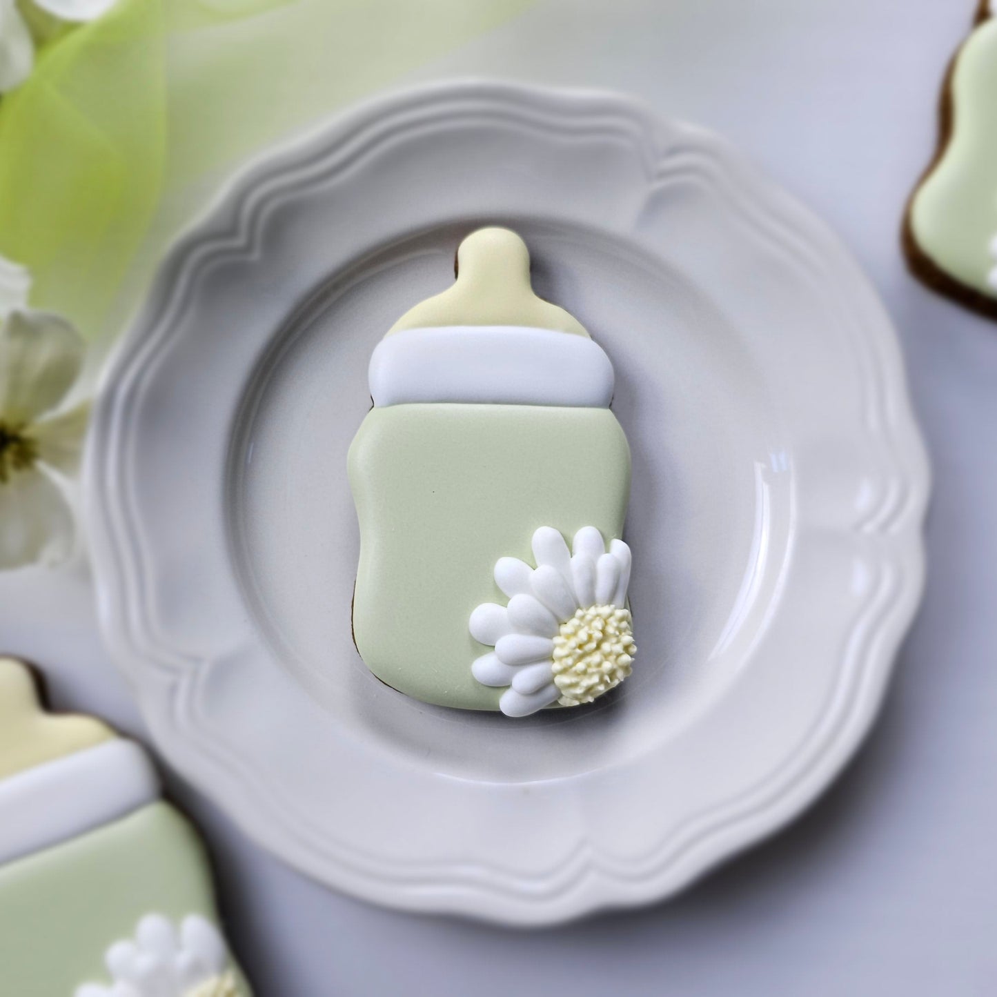 Decorative cookie shaped like baby bottle with a daisy on a plate with greenery.