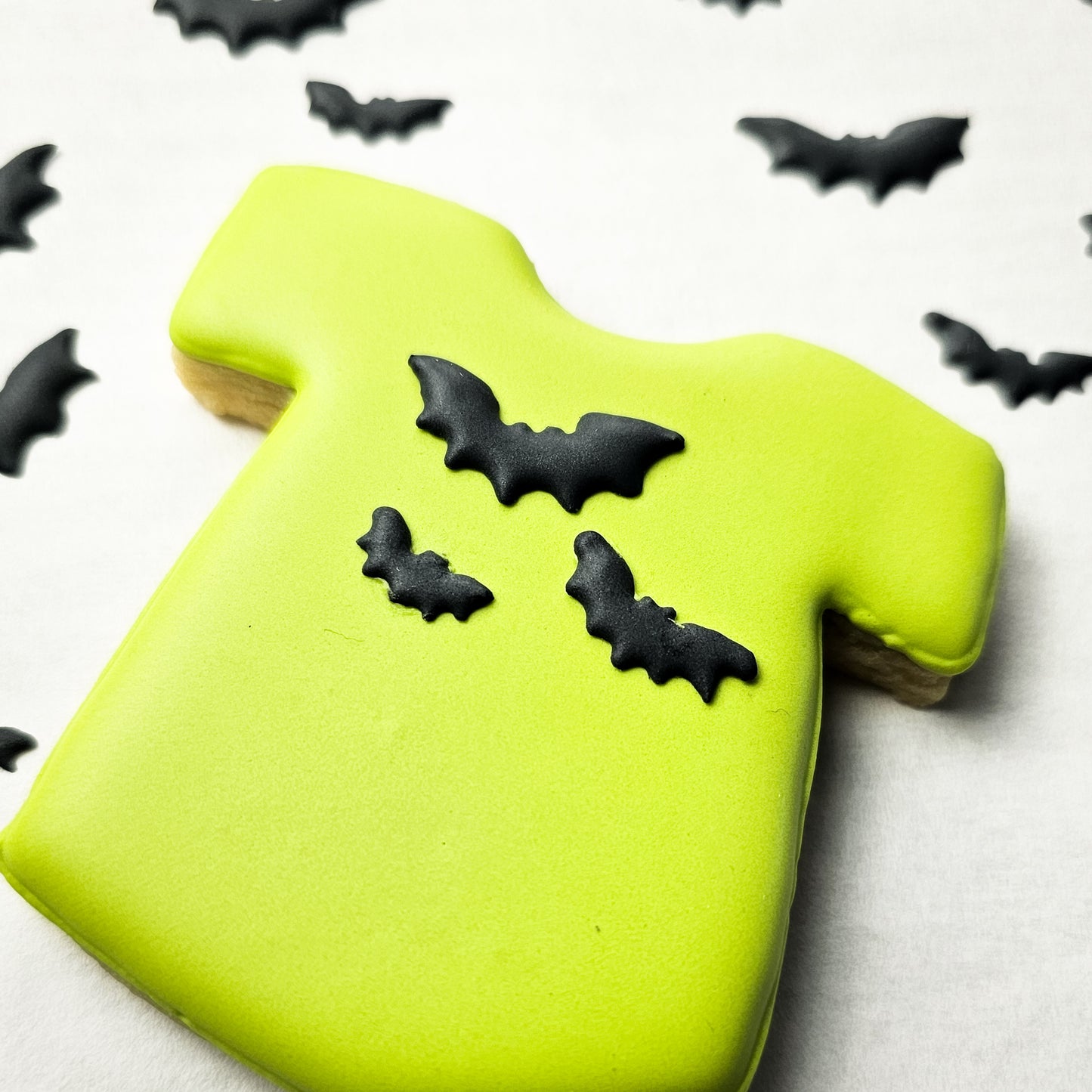 Green sugar cookie shaped like a T-shirt with black bat decorations on a white background.