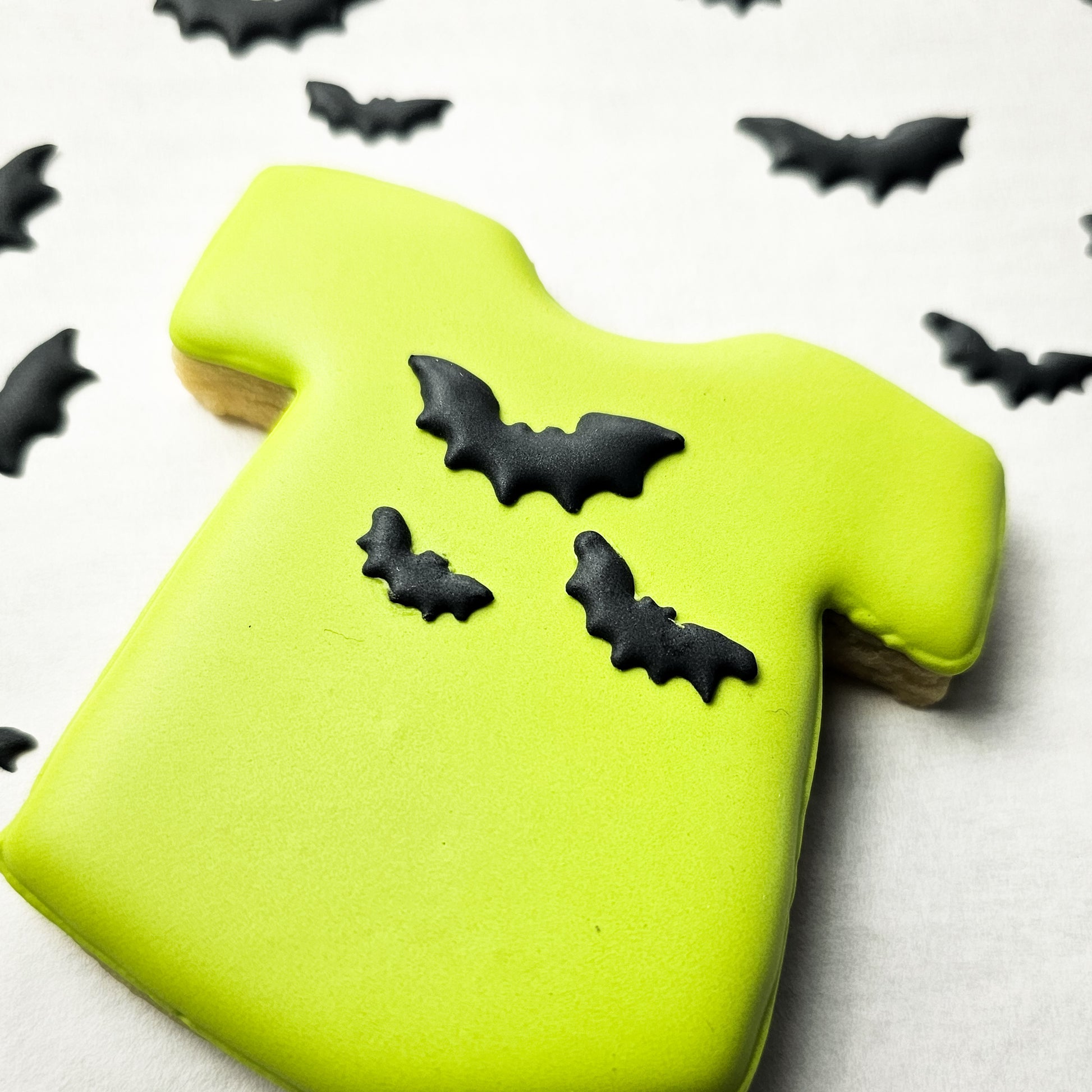 Green sugar cookie shaped like a T-shirt with black bat decorations on a white background.