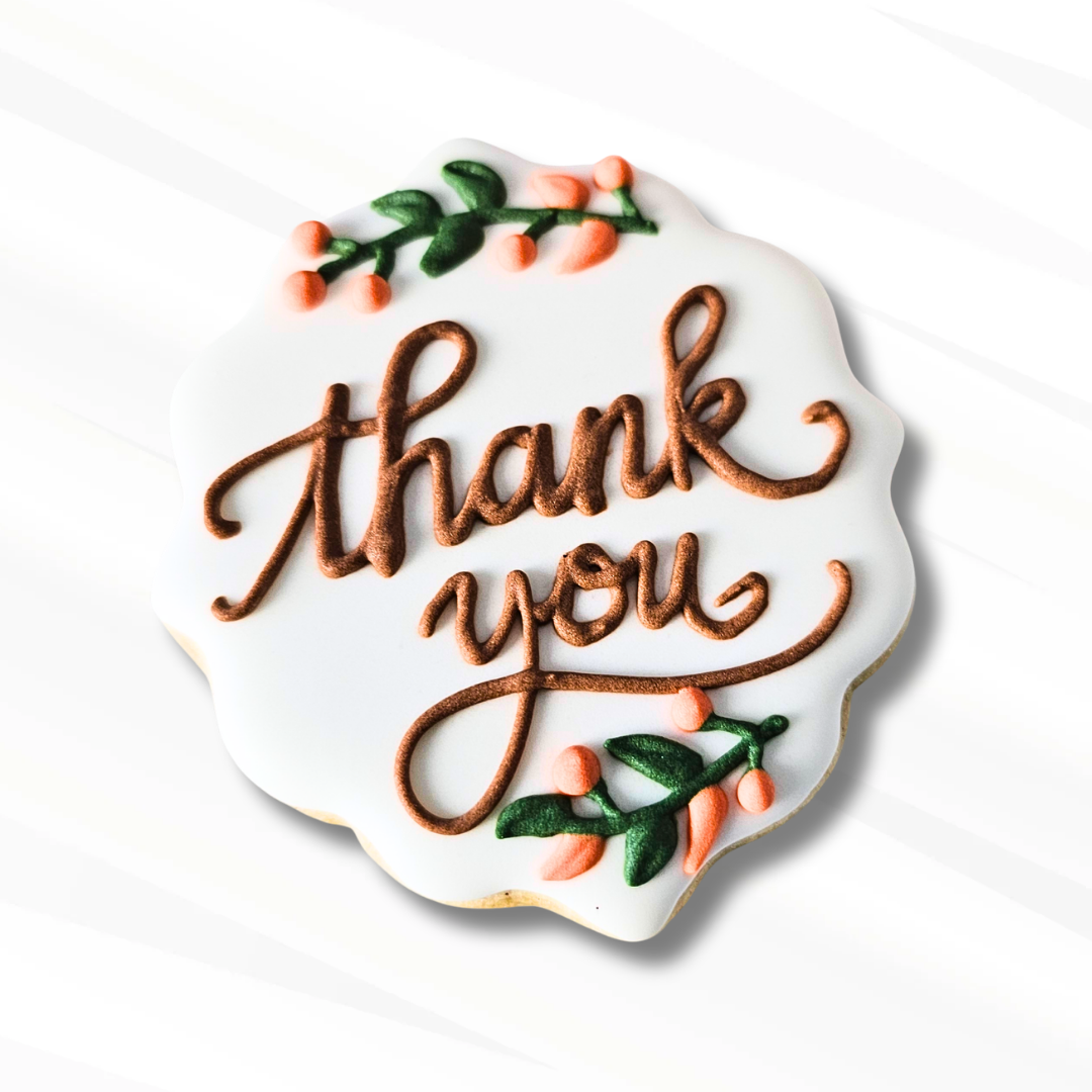 A scalloped white sugar cookie with 'thank you' written in brown icing, decorated with green and orange leaves on a white background with subtle diagonal shadows.