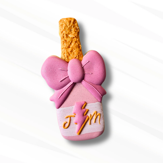 A decorative pink and orange sugar cookie shaped like a champagne bottle with a large pink bow and a lightning bolt design on the monogrammed label, set against a clean white background with subtle diagonal shadows.