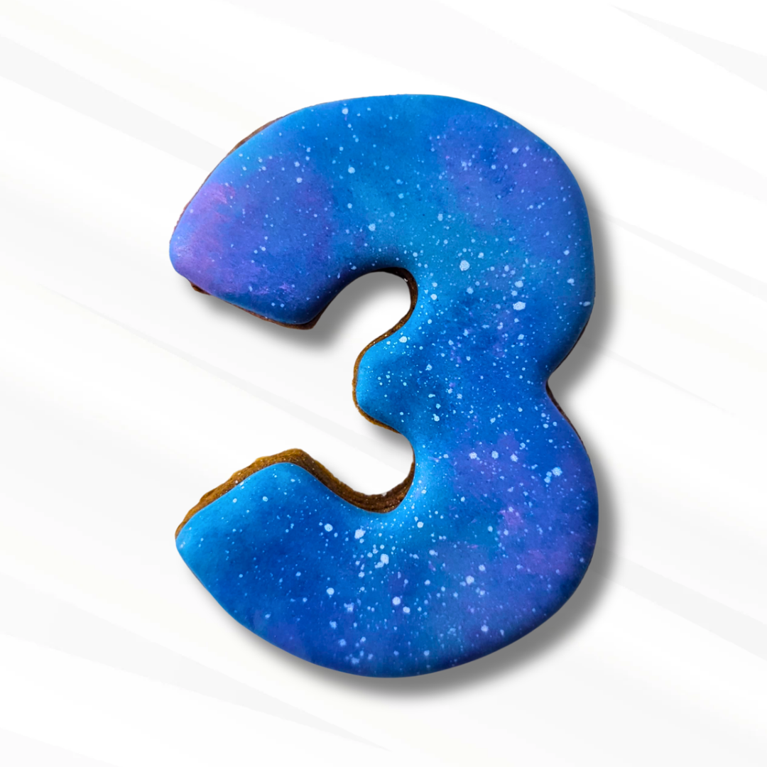 A number 3-shaped sugar cookie with blue and purple galaxy icing and white speckles, shown on a clean white background with a soft shadow.