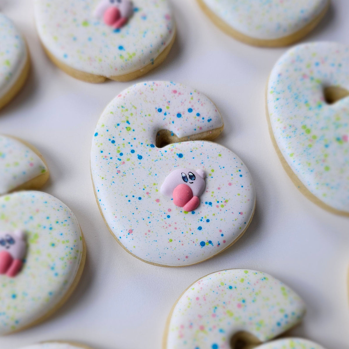 A close-up of several white-frosted number 6 sugar cookies decorated with colorful sprinkles and a small Kirby, arranged on a light background.