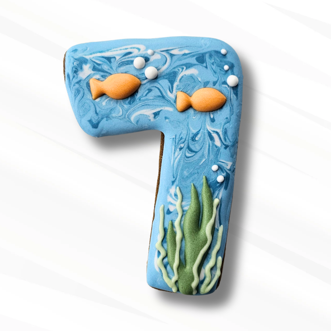 A decorated number 7 sugar cookie with an underwater theme, featuring blue marbled icing, white bubbles, green seaweed, and two orange fish, set against a clean white background.