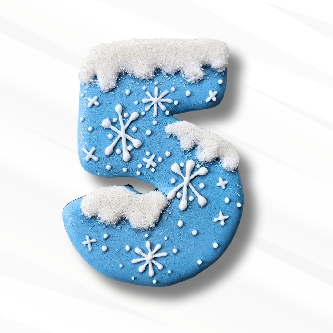 A festive number 5 shaped cookie decorated with light blue icing, white snowflake patterns, and white icing-draped sugar crystals, set against a clean white background with a soft shadow.