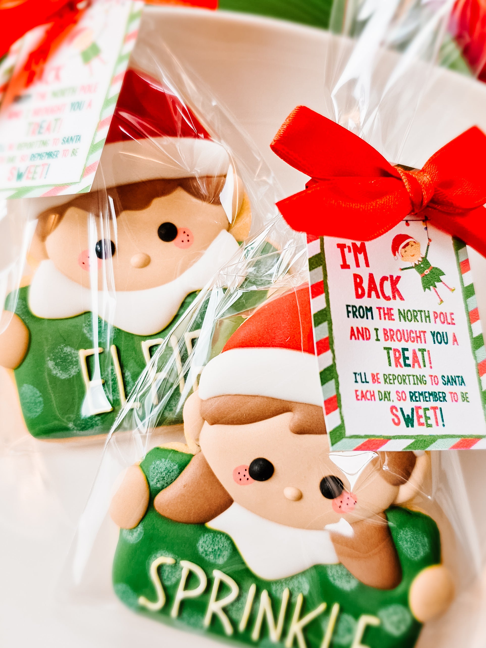 Two Christmas-themed elf sugar cookies in clear cellophane bags with red ribbons and tags.