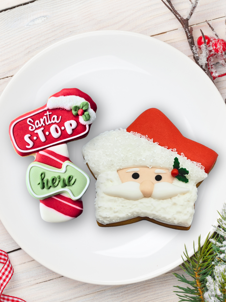 Two decorated Christmas-themed sugar cookies on a white plate: a Santa face cookie, and a red, white, and green 'Santa STOP Here' sign cookie.  The plate is set on a light wood surface with holiday decorations.