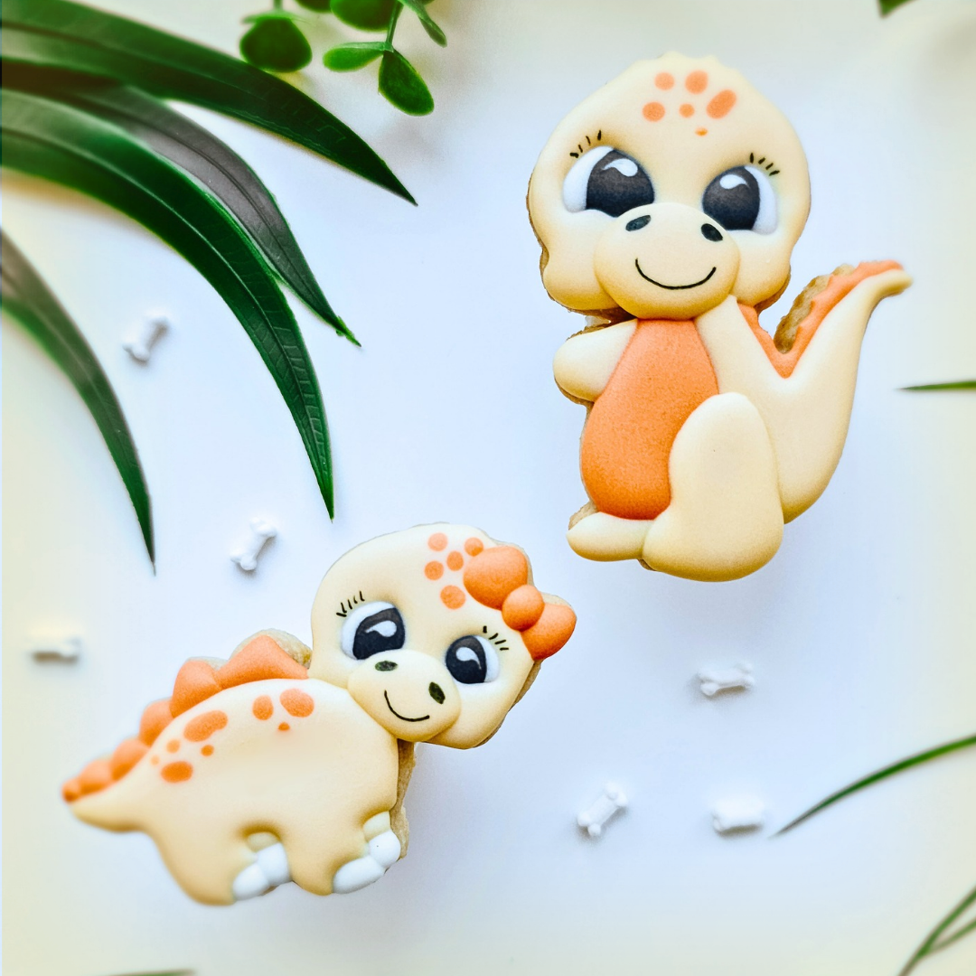Two cute dinosaur-shaped sugar cookies decorated with pastel icing, displayed on a white surface with green leaves and small white bone props.
