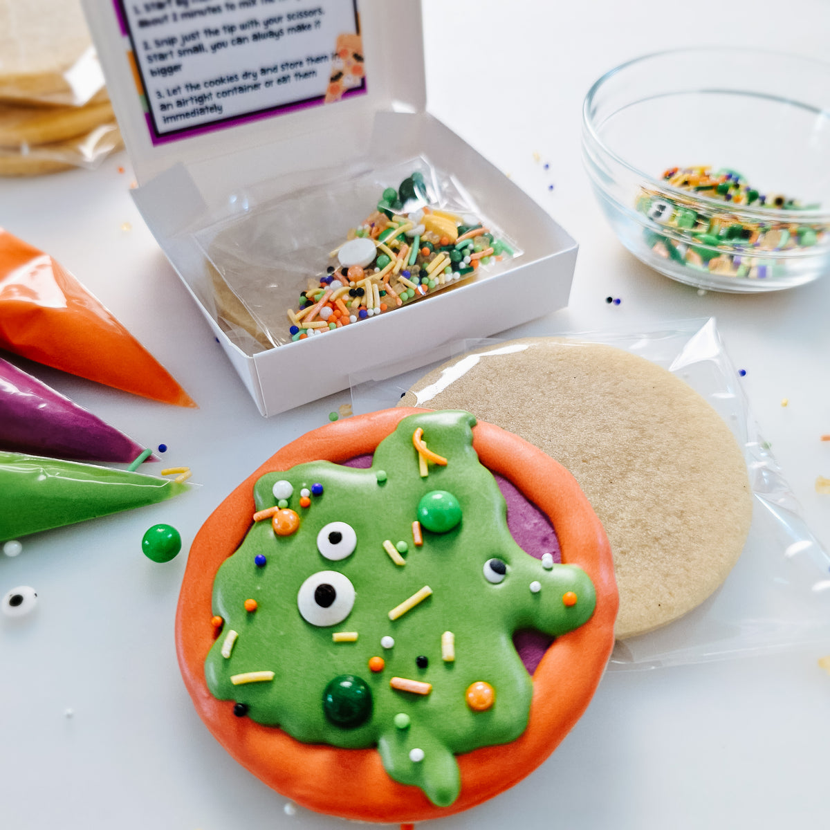 A packaged DIY cookie decorating kit on a white surface, featuring a decorated spooky pizza cookie in green, purple and orange with sprinkles, a plain cookie ready for decorating, colorful icing bags, Halloween sprinkles, and a small bowl of sprinkles.