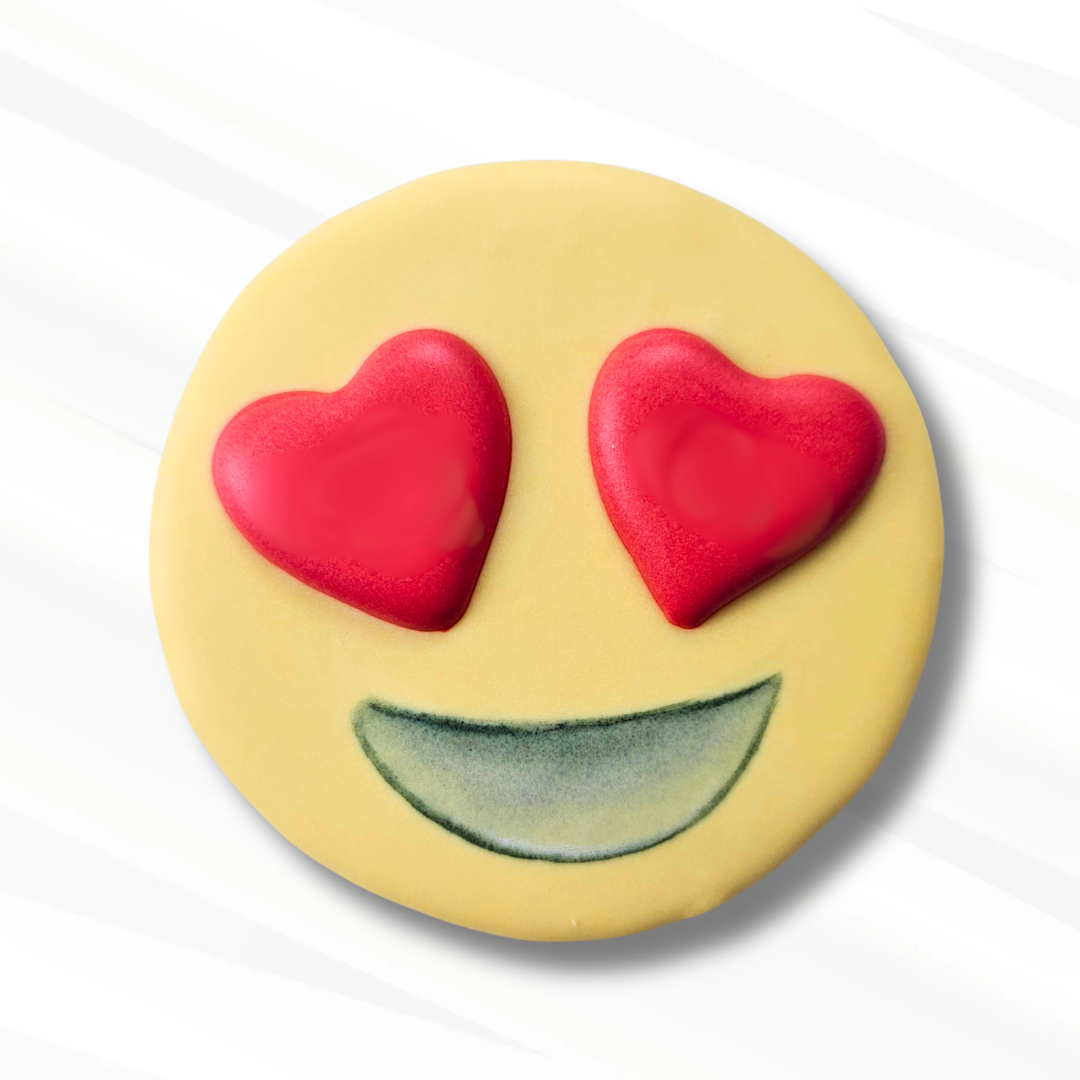 A round yellow emoji face sugar cookie with red heart-shaped eyes and a big smile, photographed on a clean white background.