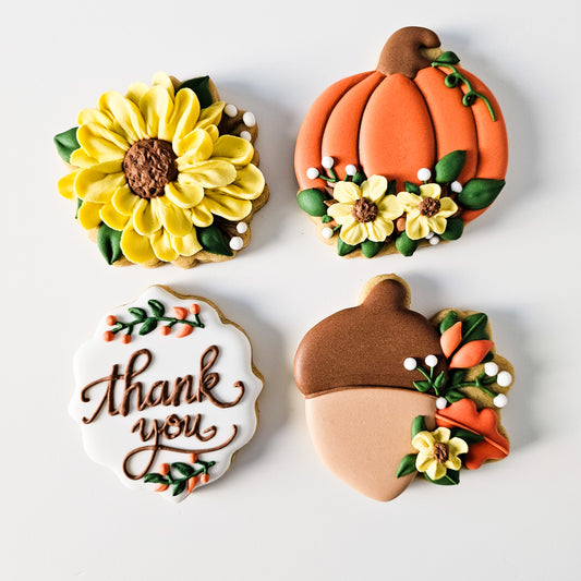 4 fall-themed decorated sugar cookies including a floral Acorn cookie, a yellow sunflower cookie, a floral pumpkin cookie, and a white plaque cookie that says thank you in brown royal icing.