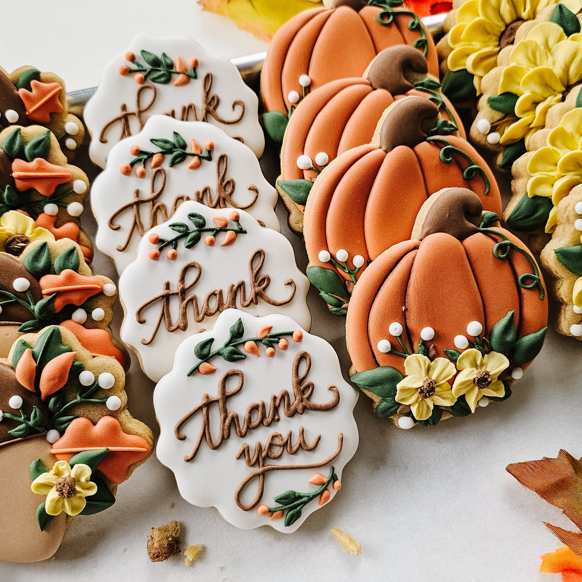 Decorated autumn-themed cookies on a white surface, including pumpkin-shaped cookies, white cookies with 'thank you' in brown script, floral Acorn cookies and sunflower cookies with yellow flowers and green leaves.