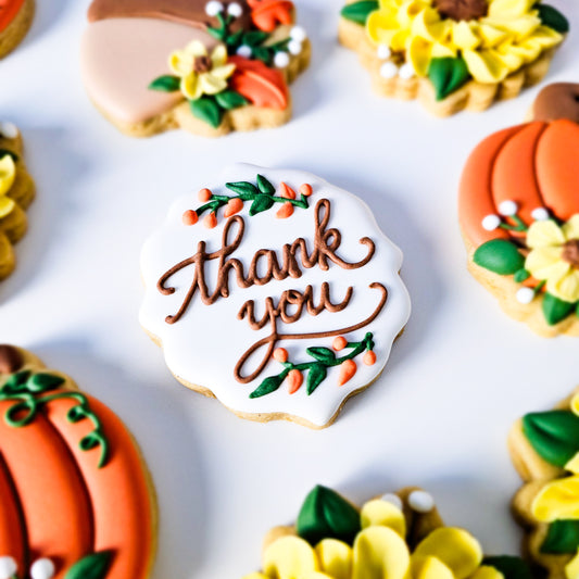 A close-up, top-down view of decorated sugar cookies on a white background. The central cookie is scalloped and white-iced with the words 'thank you' in brown script, accented with green leaves and small orange berries.