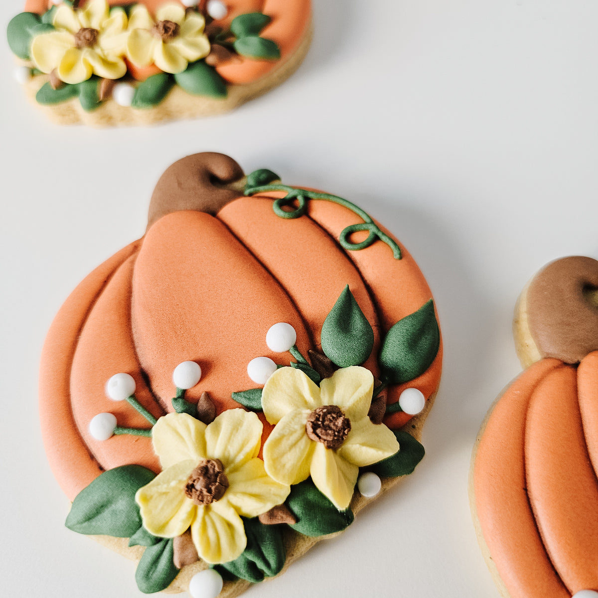 Ornate floral pumpkin sugar cookies decorated with orange icing, yellow flowers, green leaves, and small white pearl accents on a clean white background.
