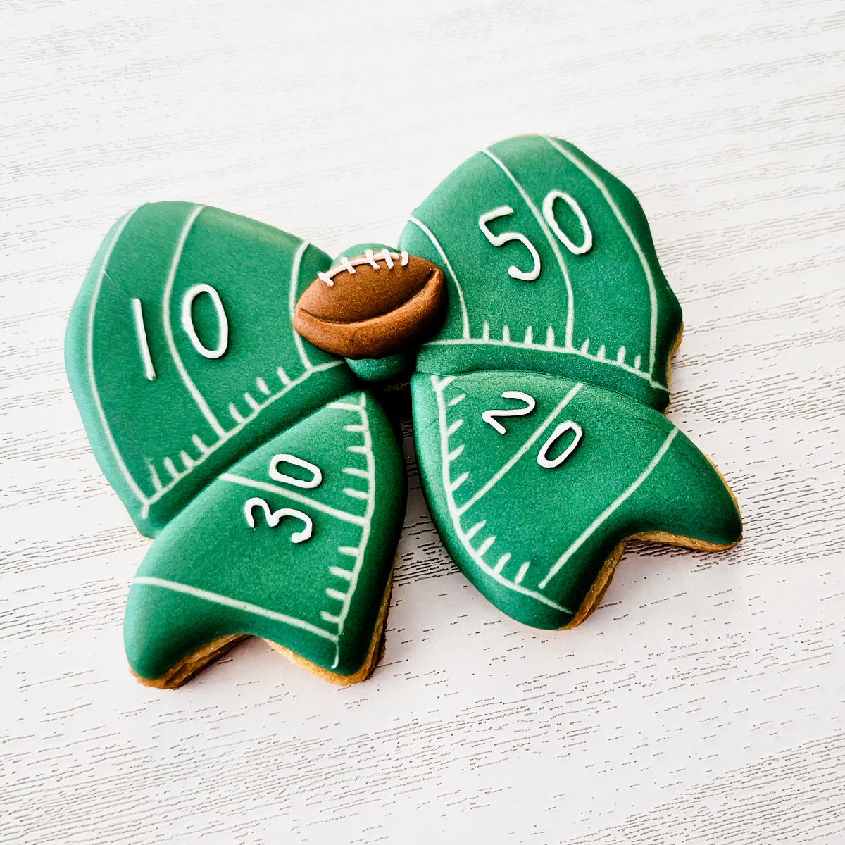 A green sugar cookie bow shaped decorated like a football field with a football royal icing transfer center on a white background