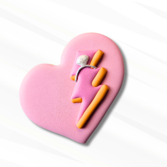 A heart-shaped pink sugar cookie featuring a lightning bolt design in pink and orange with a small silver wedding ring detail, set against a white background with soft diagonal shadows