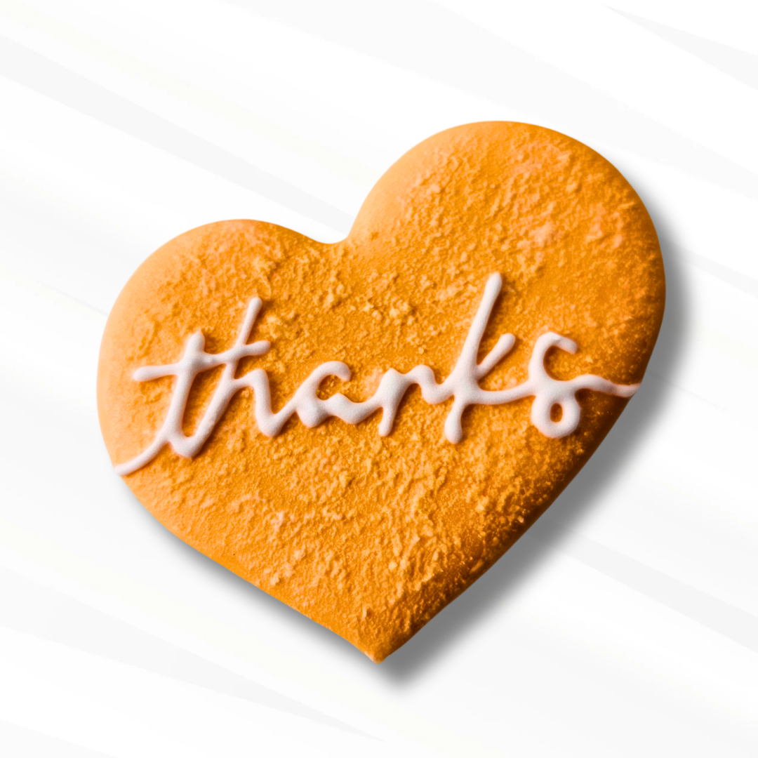 A heart-shaped orange sugar cookie with white icing writing the word 'thanks' across the front, set against a clean white background with subtle diagonal shadows.