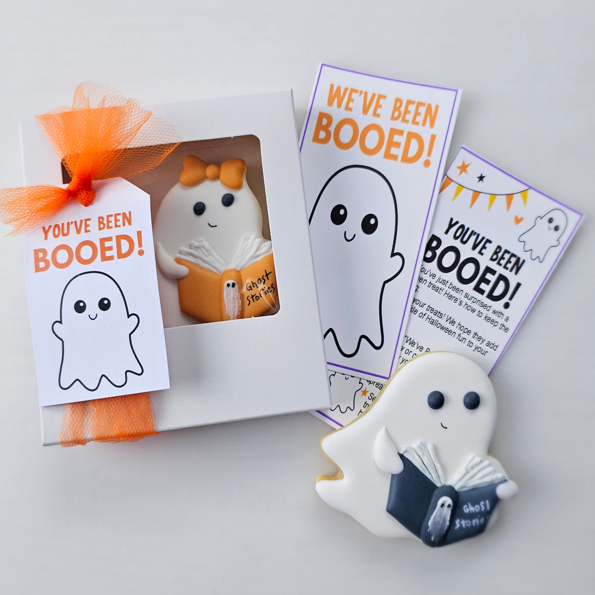 Halloween-themed sugar cookie ghost book gift set featuring a white ghost cookie holding an orange book, a white ghost-shaped tag that says 'You've been Booed' in a white gift box with a window, and printed cards that read 'YOU'VE BEEN BOOED!' and 'WE'VE BEEN BOOED!' on a light gray background.