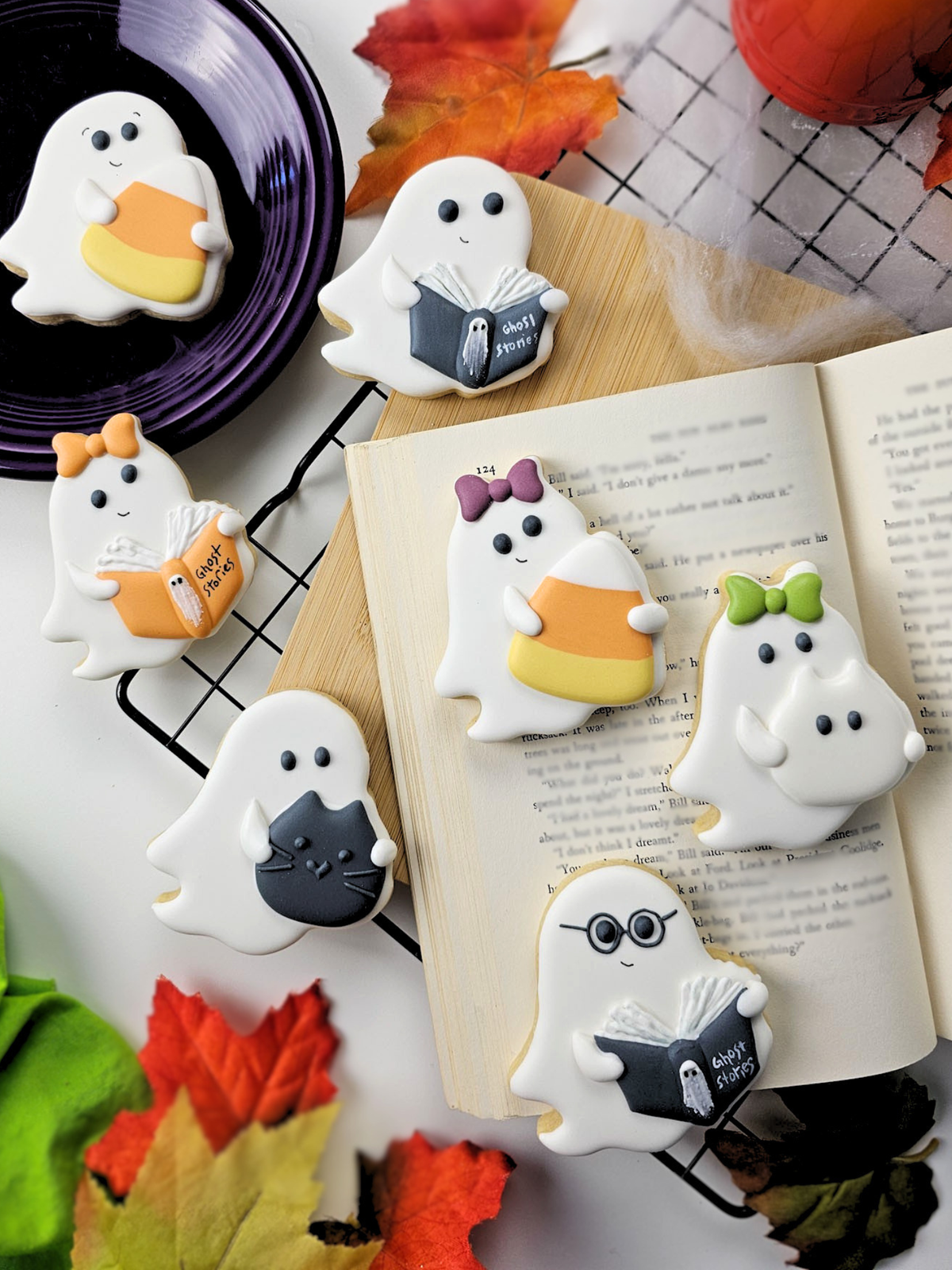 A styled flat-lay of white ghost sugar cookies decorated with Halloween-themed royal icing transfers arranged around an open book on a cooling rack with autumn leaves and a purple plate in the background.