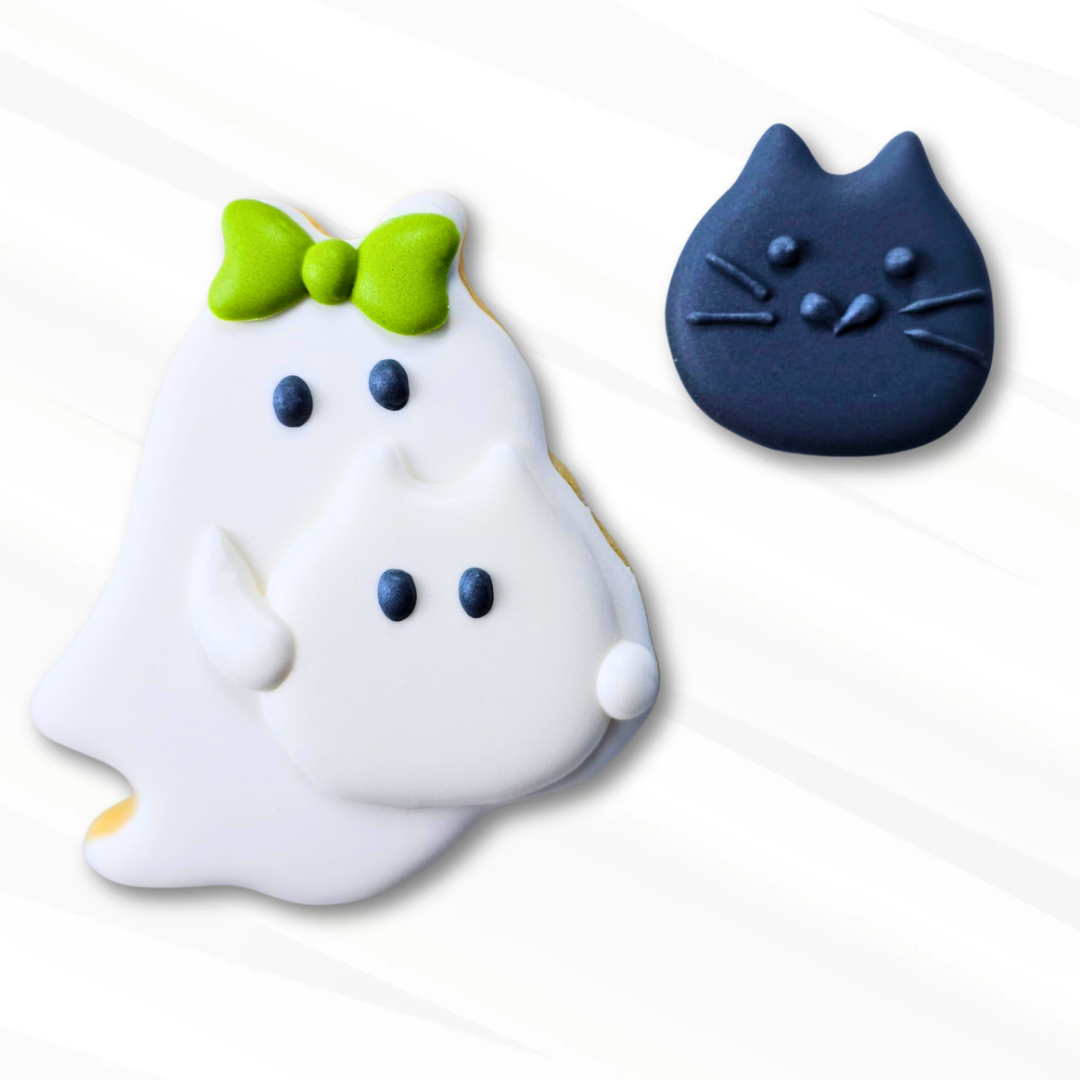 A white ghost sugar cookie decorated with a bright green bow, black eyes, and a small  Ghost Cat royal icing transfer shown next to a black cat royal icing transfer on a clean white background.