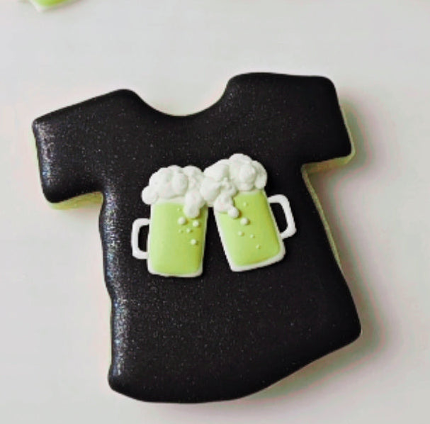 Black t-shirt-shaped cookie with green beer mug royal icing transfers on a white background