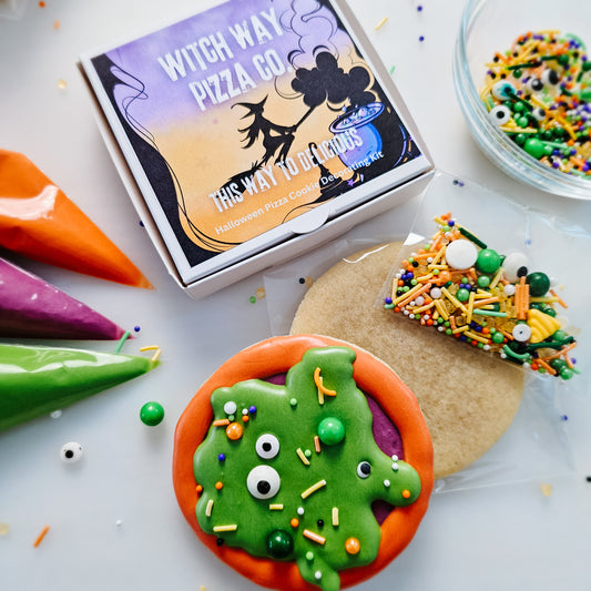 A styled flat-lay of a Halloween pizza cookie decorating kit from Witch Way Pizza Co. The picture shows a printed pizza box, a decorated gooey pizza cookie with candy eyes and sprinkles, a plain cookie, a bag of sprinkles and piping bags in orange, purple, and green on a white background.
