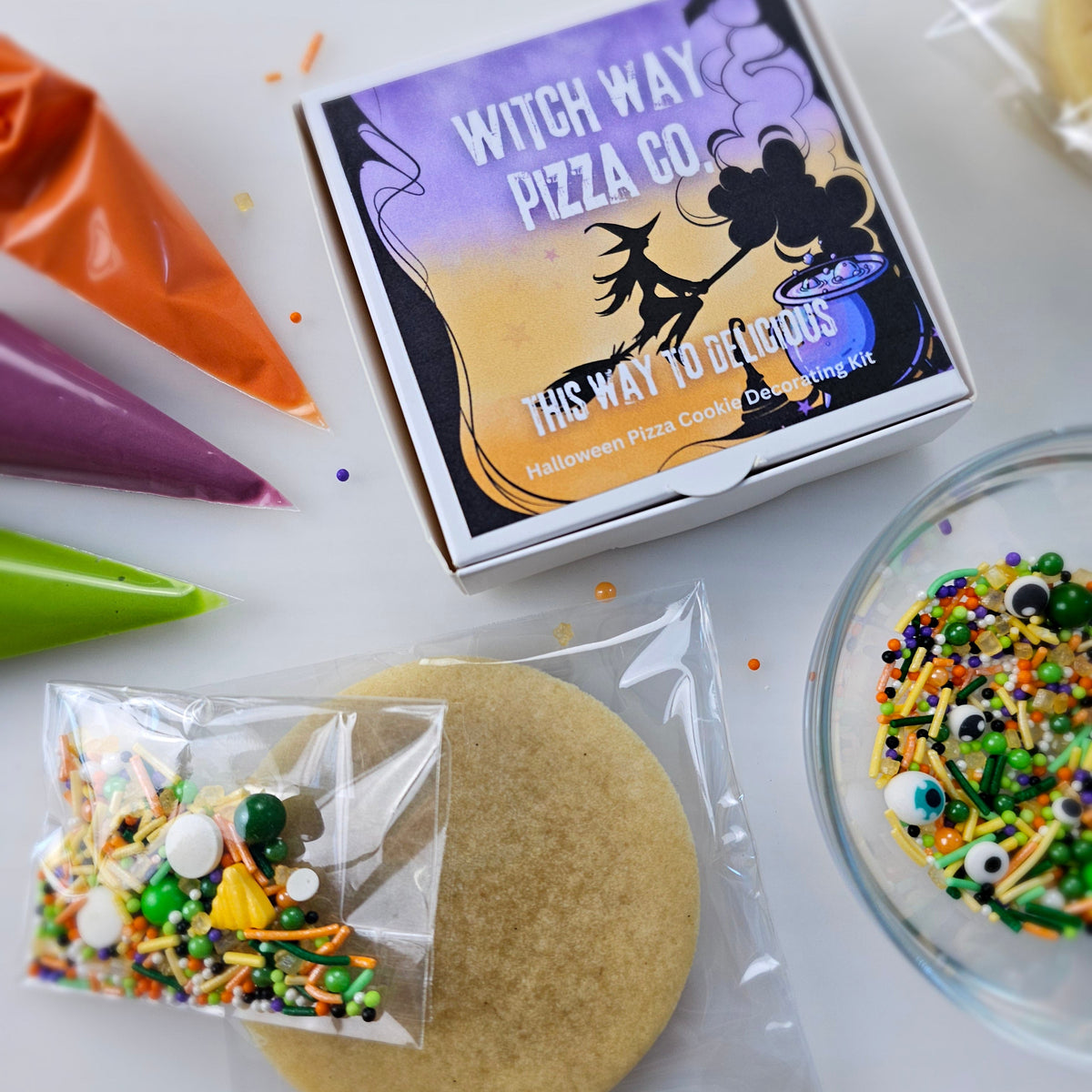 A Halloween pizza DIY sugar cookie kit from Witch Way Pizza Co. featuring a pizza box, a plain round cookie, a small bag of sprinkles, and piping bags with orange, purple, and green icing on a white surface.