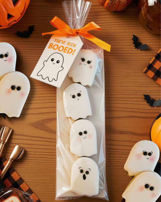 A package of ghost-shaped cookies on a wooden table surrounded by Halloween decorations. The cookies are white with cute black eyes, pink cheeks, and small smiley faces.