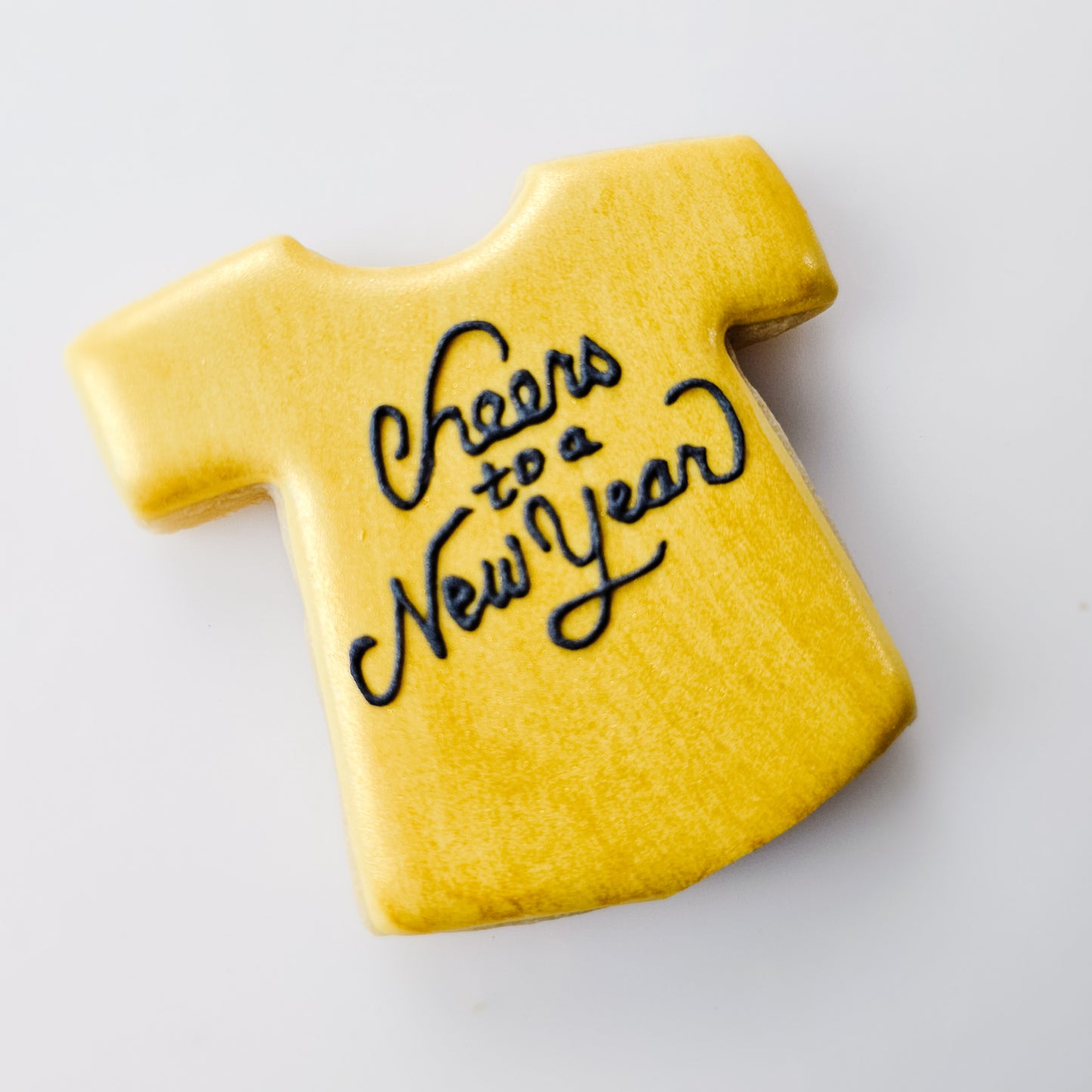 Gold sugar cookie shaped like a t-shirt with 'Cheers to a New Year' text on a white background