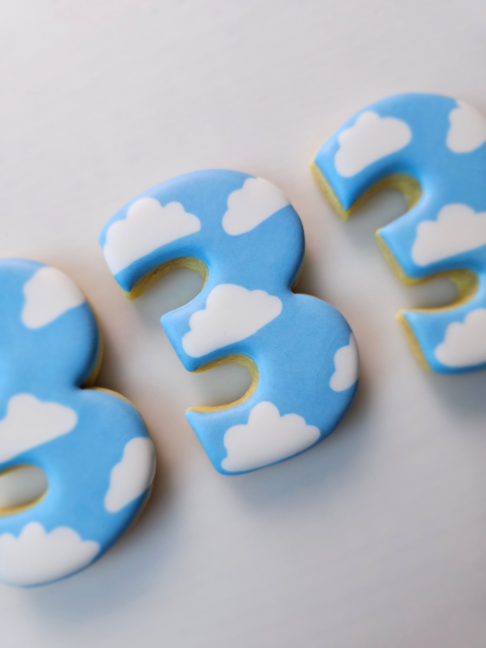 Decorative number 3 sugar cookies iced with clouds and blue sky on a light gray background