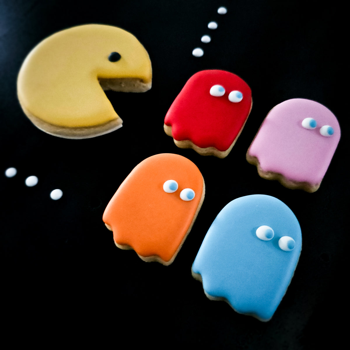 Pac-Man inspired sugar cookies in yellow, red, orange, pink, and blue, arranged on a black background with white dots.