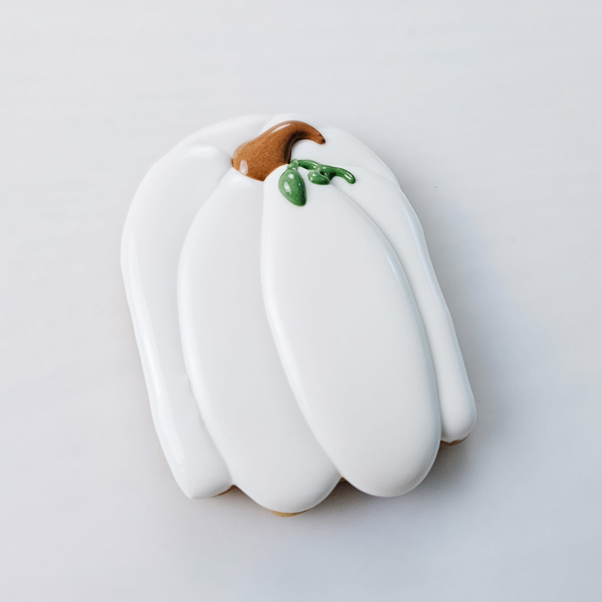 A white pumpkin-shaped cookie with smooth icing, featuring a brown stem and green leaf details, photographed on a clean white background.