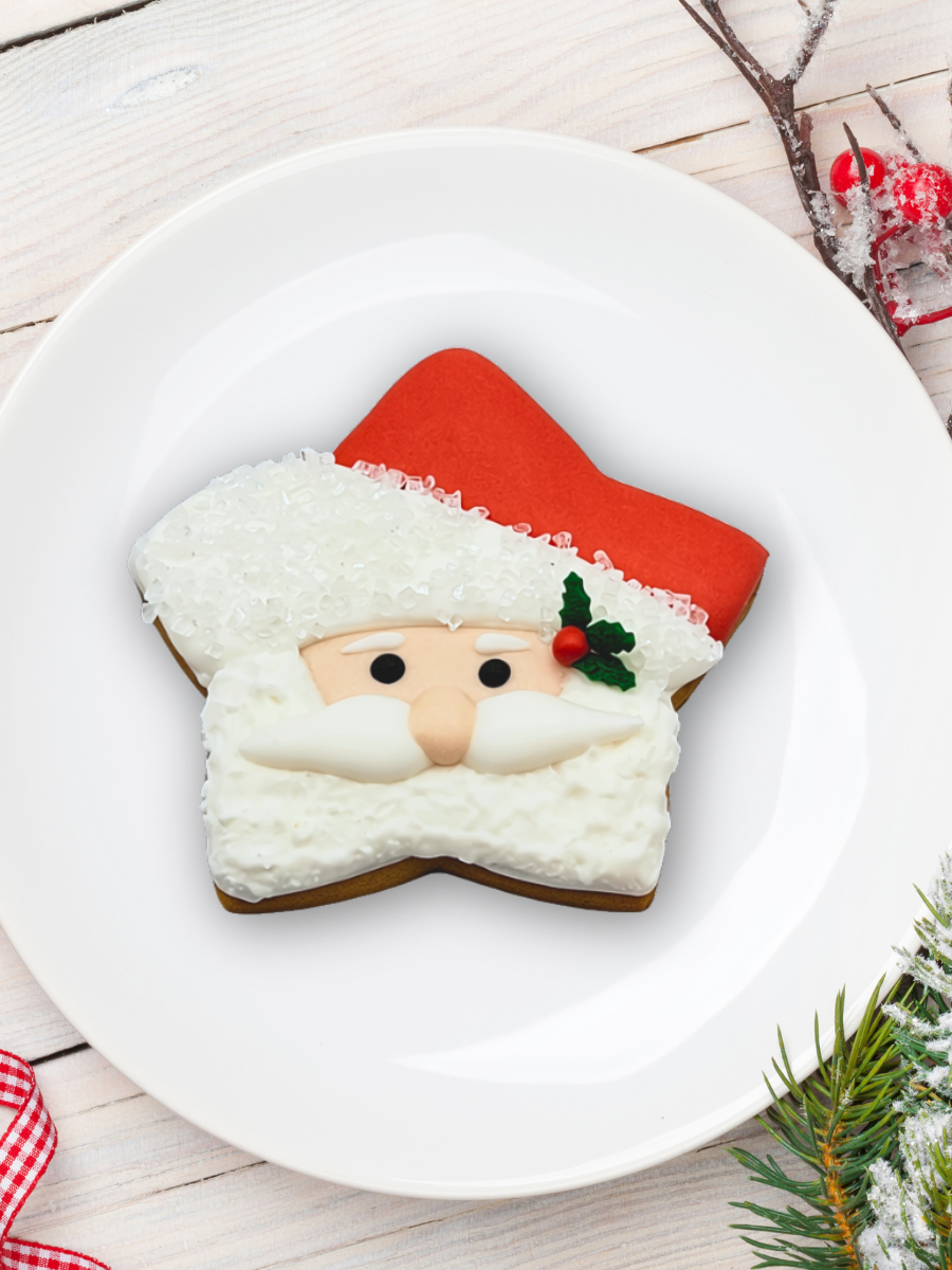 A Santa Claus Star shaped sugar cookie with red hat, white beard, and small holly detail, served on a white plate over a light wood surface with holiday decorations.