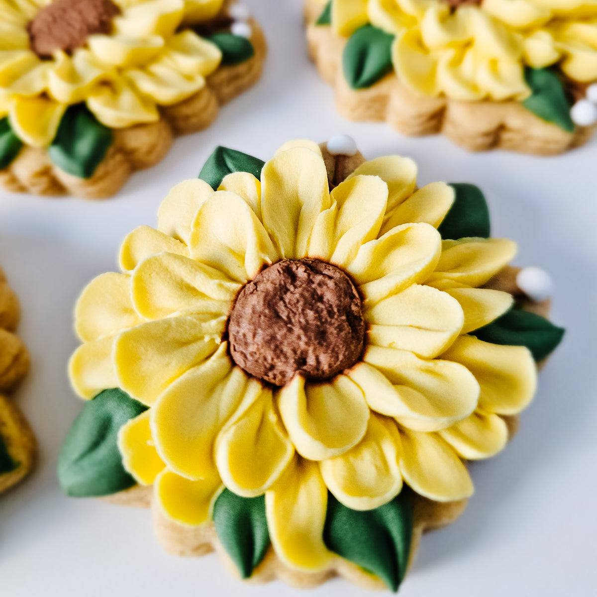 A close-up of a decorated sunflower-shaped sugar cookies with yellow piped petals, a brown textured center, and green leaf accents on a white background.