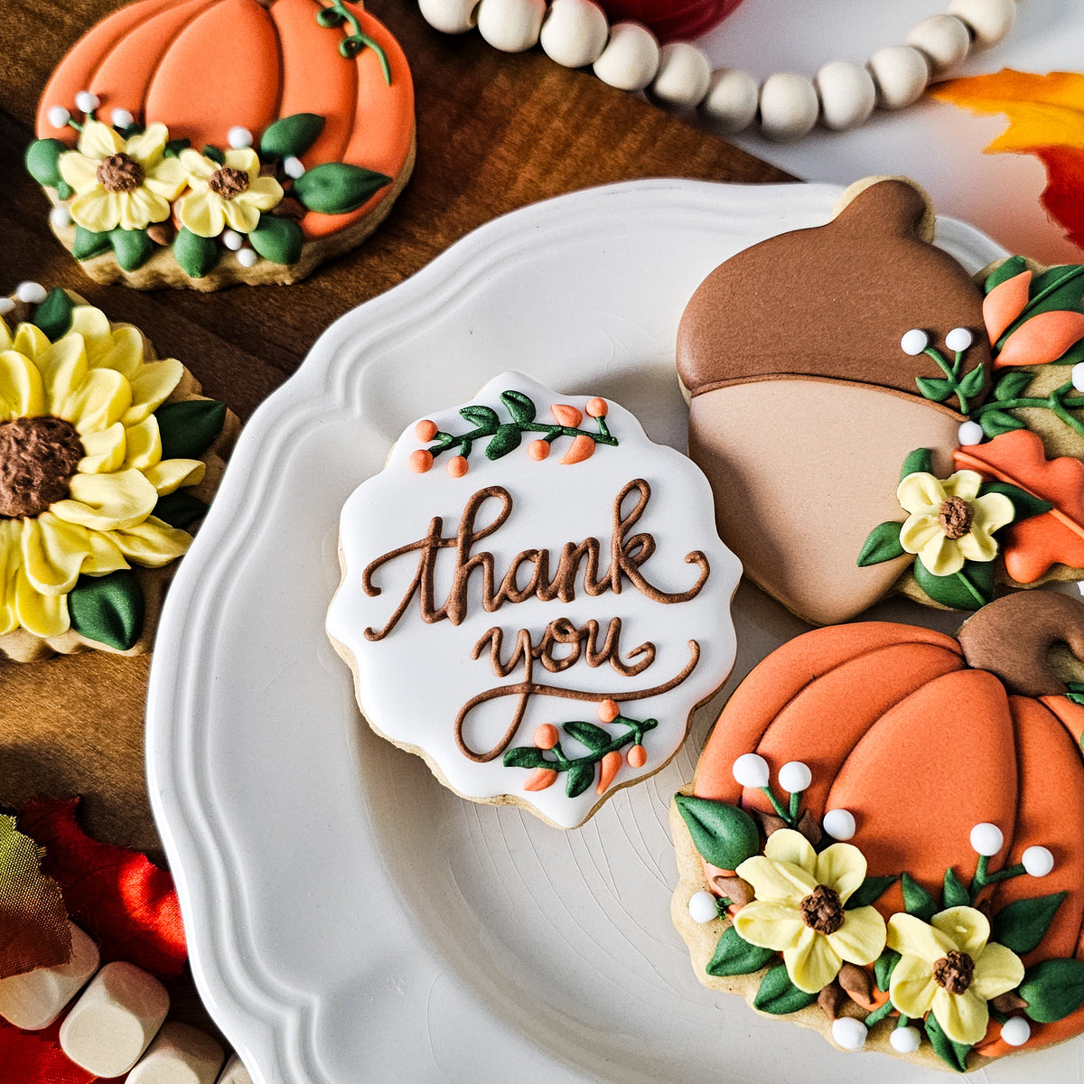 Decorated fall-themed sugar cookies on a white plate, including a pumpkin cookie with yellow flowers, an acorn cookie, and a scalloped white cookie with 'thank you' in brown script.