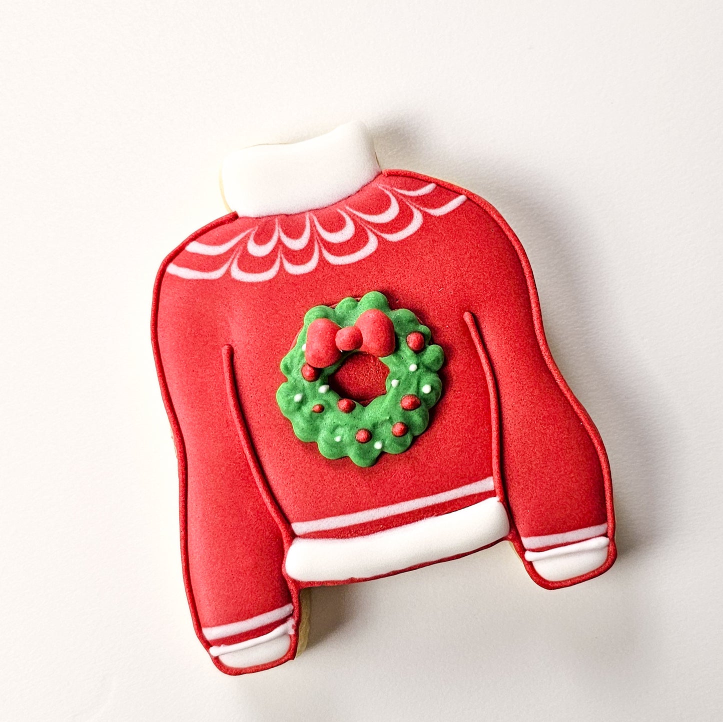 Red sweater-shaped sugar cookie with a green wreath and red bow on a white background