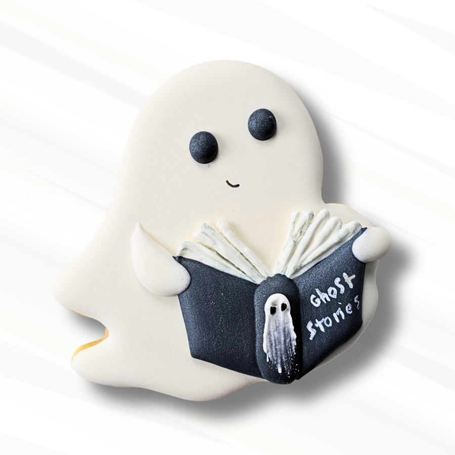 A ghost-shaped sugar cookie with a white body, dark eyes, a small smile, and a dark book reading 'Ghost Stories'. The cookie is photographed on a clean white background with soft shadows.