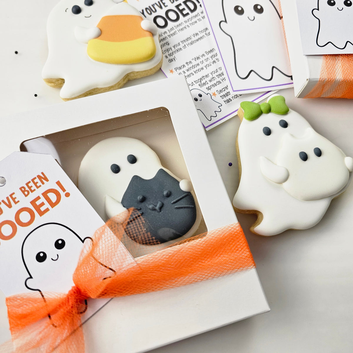 Halloween ghost-shaped sugar cookies holding a black cat royal icing transfer shown in a gift box with an orange ribbon and a tag that reads 'YOU'VE BEEN BOOED!'.