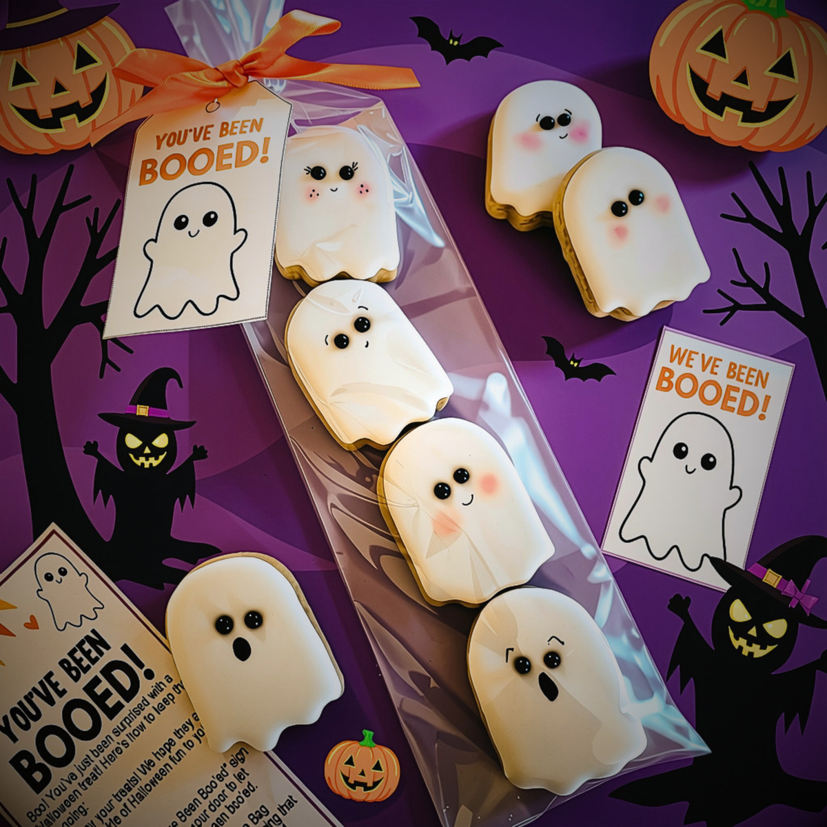 Halloween ghost-shaped cookies decorated with white icing and cute faces, packaged in a clear bag with a 'You've Been Booed!' tag. The scene is styled on a purple background with pumpkins, bats, and witch silhouettes.