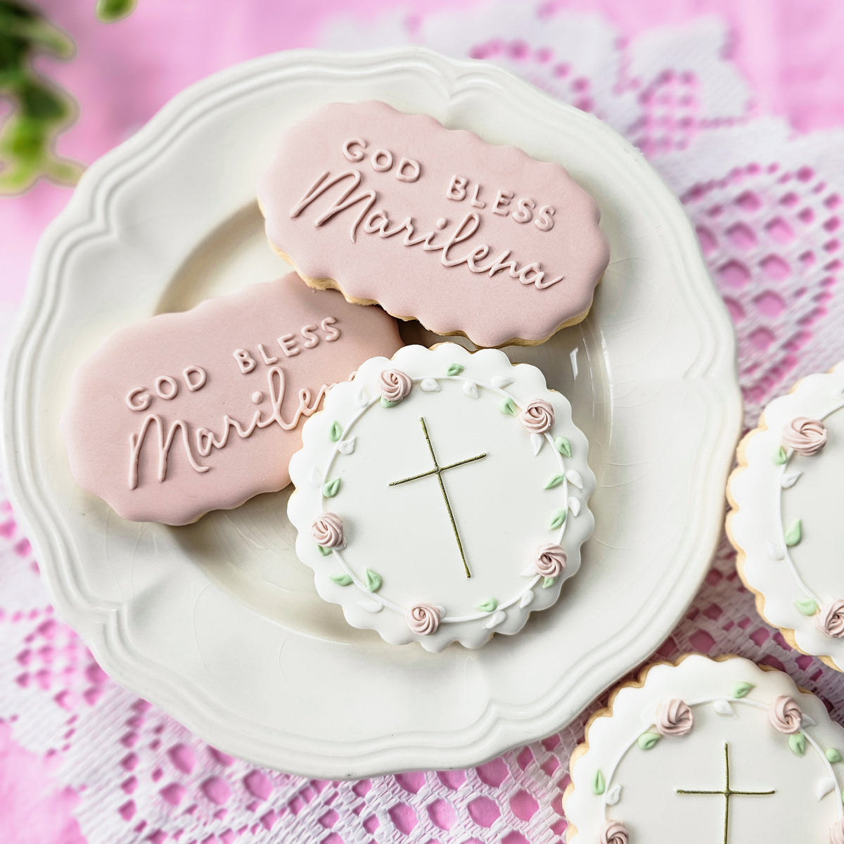 Decorated sugar cookies on a white plate, including pink oval-shaped cookies with 'God Bless Marilena' and a round white cookie with a gold cross and floral border.